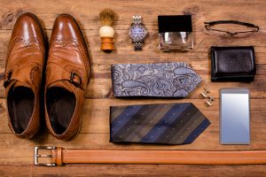 Variety of men's accessories organized in knolling arrangement.  Items include: dress shoes, ties, glasses, wallet, watch, shaving brush, cologne, belt, smart phone.  The items all lie on a wooden desk or table.   Men's personal accessories, clothing themes.  Father's Day.  Fashion.  Business.  Retail.